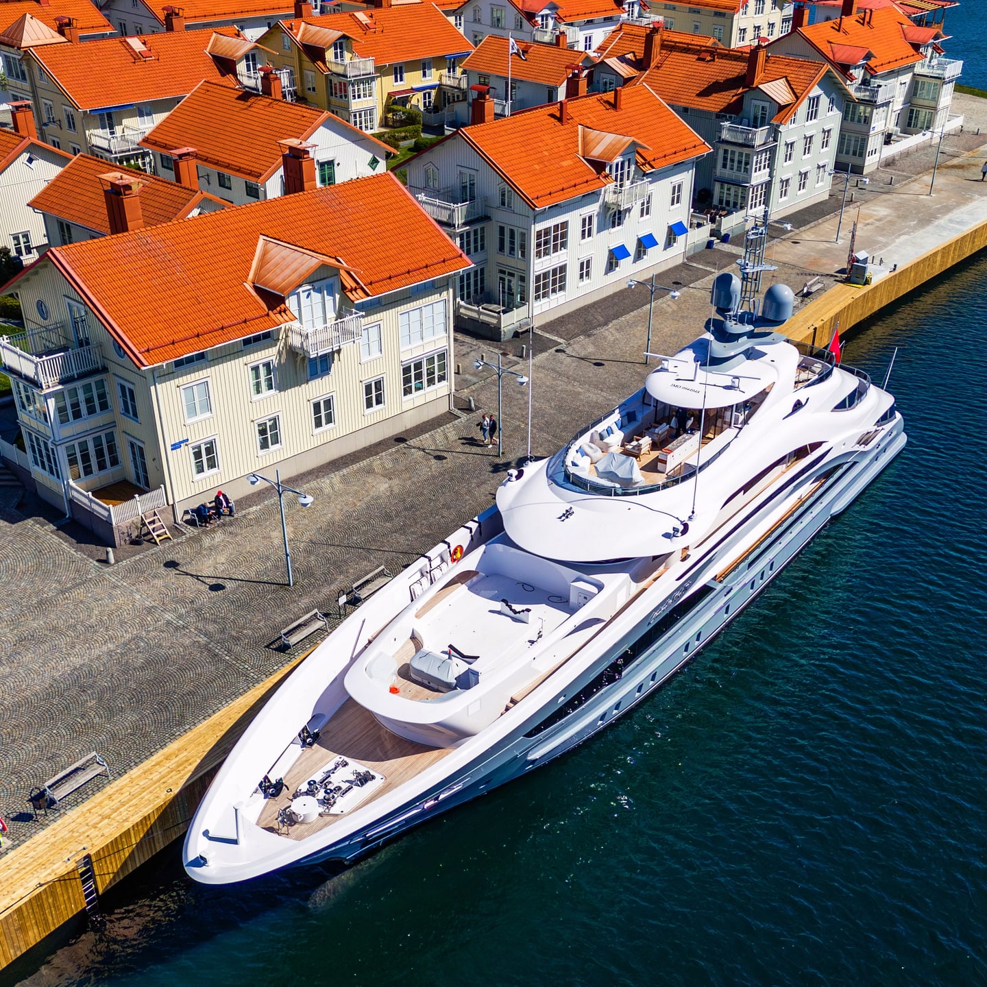 A white yacht tied up along a quay below red-roofed waterfront buildings.